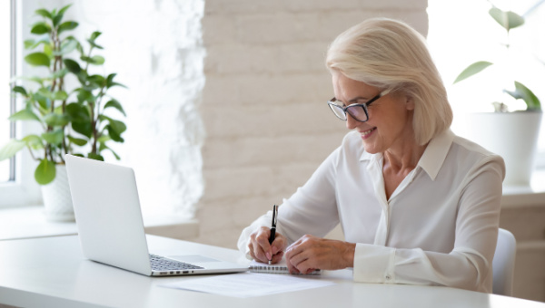 Senior woman in a well lit room taking notes in front of a laptop
