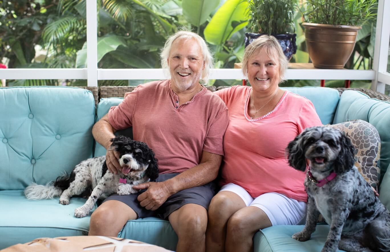 An elderly man and woman sitting on their couch smiling with their two dogs