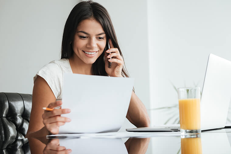 A seated woman reading a piece of paper next to a laptop computer on a table