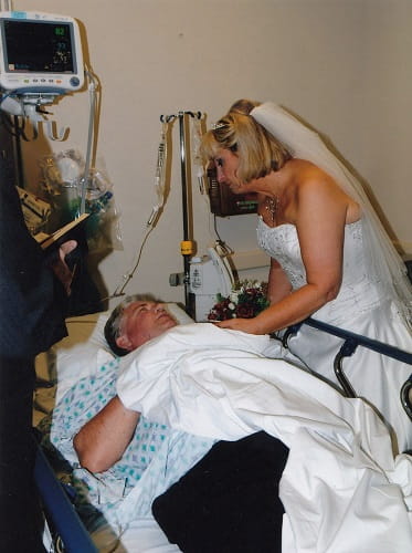 Dianna stands over Les in his hospital bed and comforts him