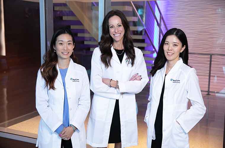 Three female primary care providers stand together wearing white coats and smiling: Minjoo Kim, DO, Jenna Fluegge, MD, and Jinjoo Hong, APRN.