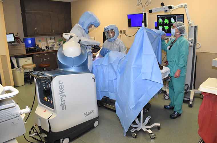 Dr. Grayson guides the robotic arm in a simulation of a robotic surgery knee replacement. Dr. Grayson and two other team members are all wearing surgical garb.