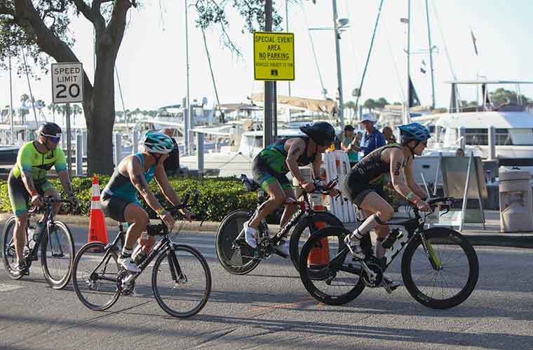Four bicyclers wearing athletic outfits ride by city waterfront in athletic competition. There are boats in the background. 