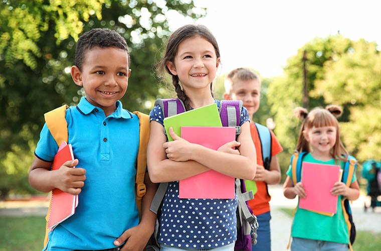 Four children who are elementary school age holding folders and wearing backpacks outside a school.