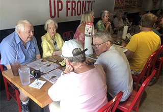 A gray-haired man in a blue shirt sits next to a gray-haired woman in a yellow jacket. They are across from a woman in a pink shirt and a man with glasses. They are talking and looking at menus at a restaurant. Other people are also at the table talking to each other.