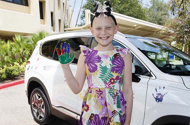 Young girl with cancer smiles while showing up her hand that is dipped in red, blue and green paint moments before she places it on a white vehicle.