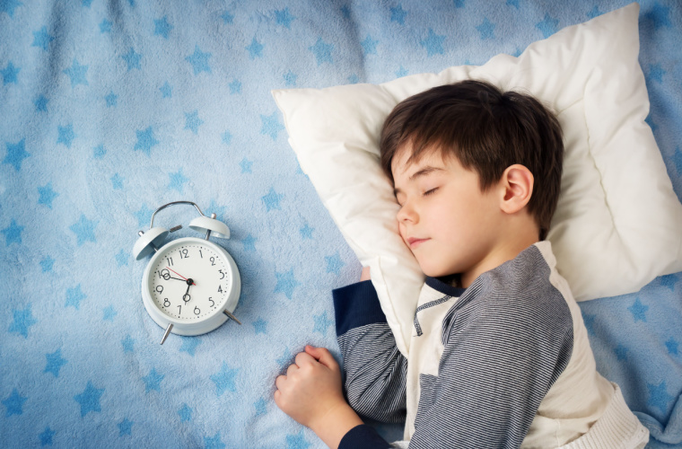 Little boy with brown hair is sleeping in bed with an alarm clock.