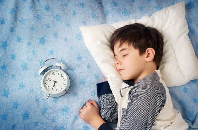 Little boy with brown hair is sleeping in bed with an alarm clock.