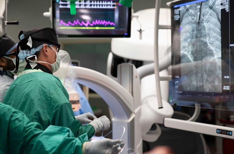 Two male cardiovascular surgeons in scrubs perform an image-guided, minimally invasive Transcatheter Aortic Value Replacement (TAVR) procedure in an operating room while viewing an image of a heart on a screen.