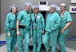 Five members of the surgical team, all wearing green scrubs and surgical caps, stand by the new robotic system.