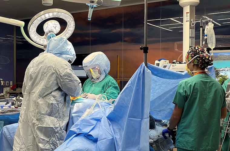 Three people in surgical gear standing over an operating room table. The patient is covered with a surgical awning with the back of her head in the photo. A large light is overhead.
