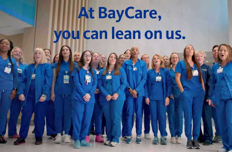 A large group of BayCare nurses wearing royal blue scrubs sing during filming of a new television commercial.  The words "At BayCare, you can lean on us." are superimposed on the screen. 