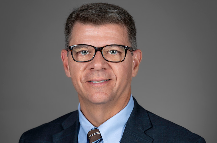 Executive portrait of C. Todd Jones with a gray background. He wears eyeglasses, a dark suite, blue shirt and striped tie.