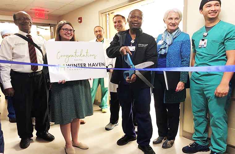 A Winter Haven Hospital Team Member uses giant scissors to cut a blue ribbon noting the cath lab opening while he is surrounded by hospital colleagues and members of the local Chamber of Commerce