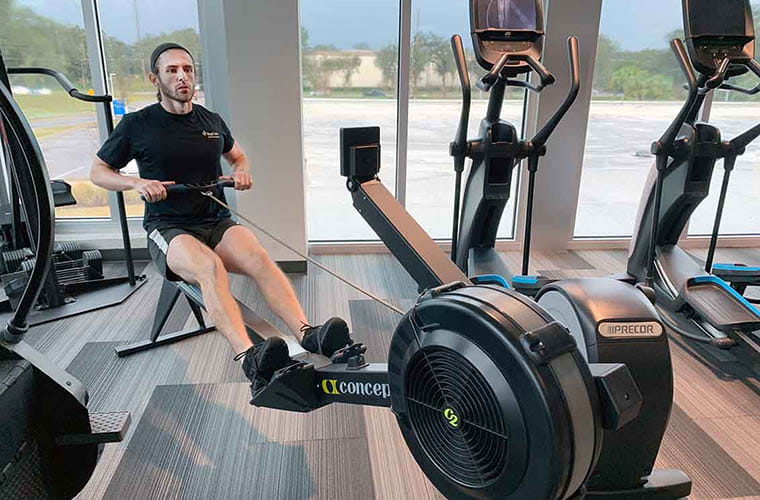 A fitness trainer, wearing a black t-shirt and black shorts, pulls back as he is working out on a rowing machine inside a fitness center, with additional fitness equipment surrounding him.