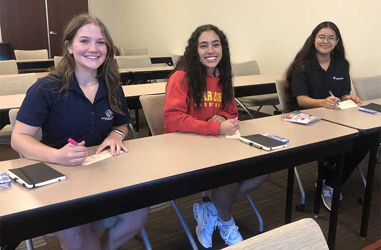 Three smiling teenage girls sit together in a classroom during their first day of the new training program.