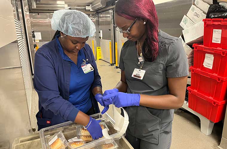 Two young women wearing scrubs, hair nets and gloves put together a food item for a patient tray in a hospital kitchen.