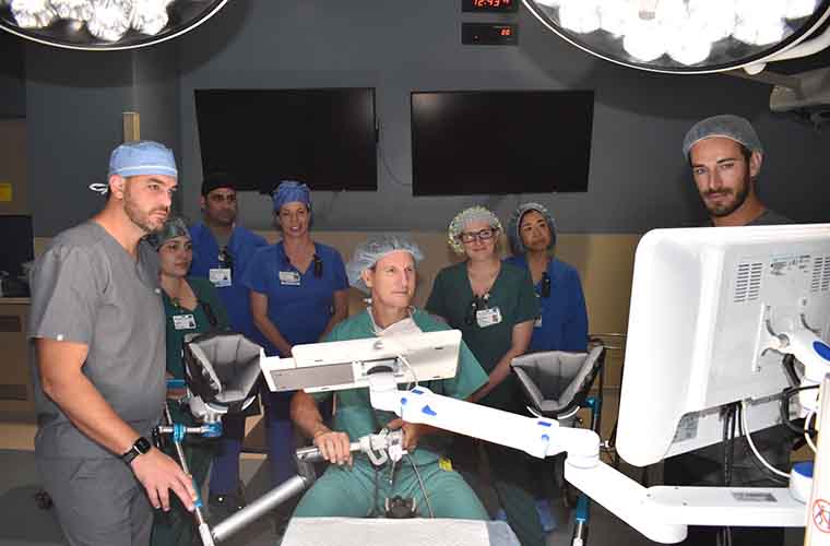 The photo simulates a BPH Aquablation surgery with the surgeon seated in back of the surgical table and looking at an imaging monitor. The surgeon's face is visible from this angle. Seven surgical team members who assist are looking on while surrounding the surgeon and the surgical table. 