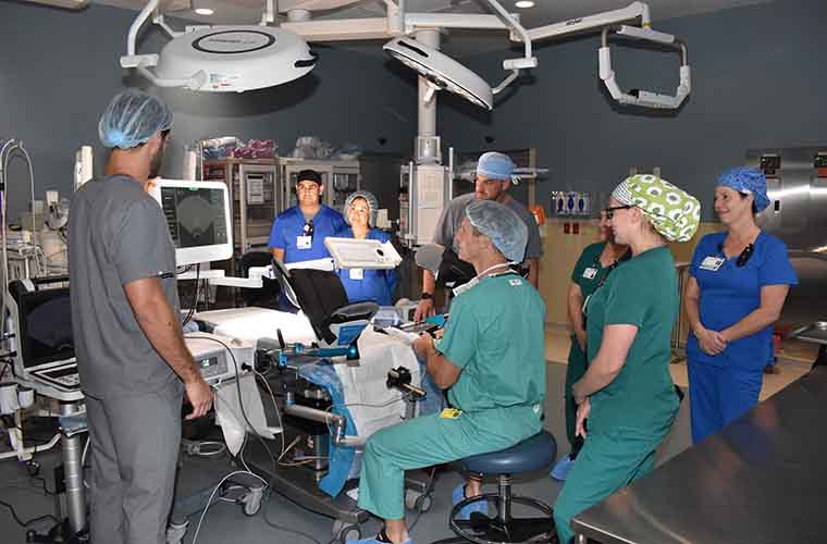 The photo simulates a BPH Aquablation surgery. The surgeon is seated at the console facing a monitor. The side of his face and back are visible from this angle. The surgeon guides the robotic Aquablation system using imaging on a monitor. Seven surgical team members look on while surrounding the surgeon and the surgical table.