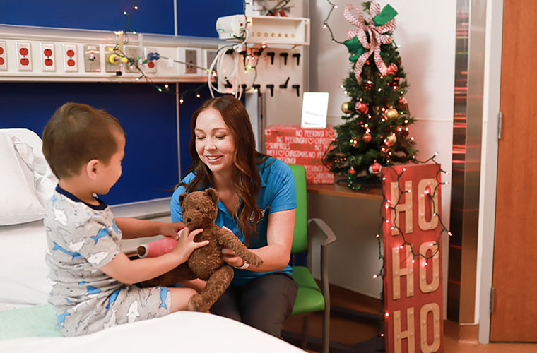 Toddler boy on hospital bed holds brown stuffed bear while playing with a woman who is one of his clinicians. The hospital room is decorated with a Christmas tree.