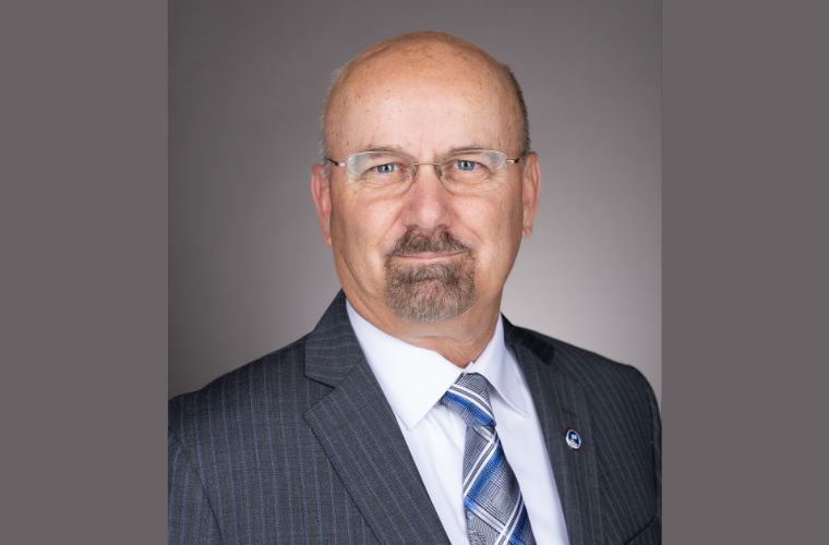 Headshot of John J. Loetscher wearing a gray pinstripe suit, a white button up shirt with a gray, blue and white vertical striped tie and a lapel on his left side. He is wearing small glasses, has a goatee and is bald.