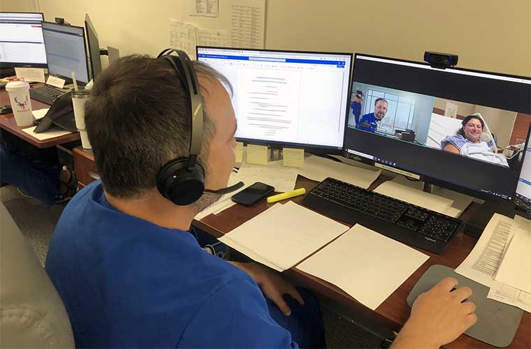 Nurse in blue scrubs sitting at desk with multiple monitors talks to a patient on his screen.