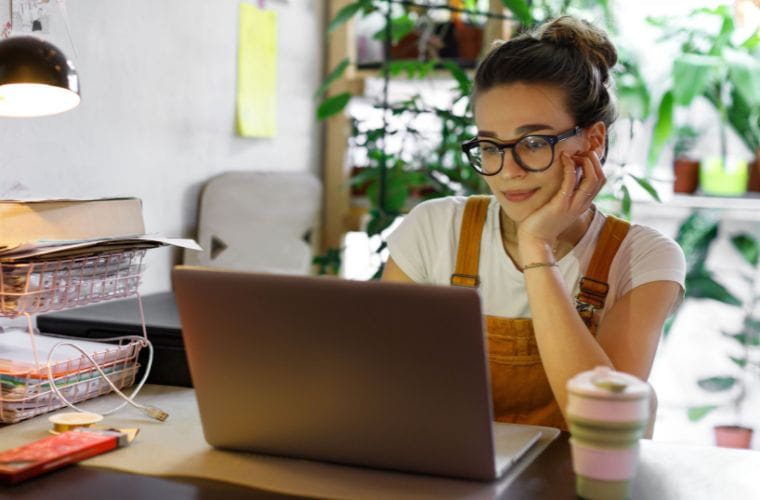 A young woman with brown hair in a bun wearing glasses, a white T-shirt and orange overalls sits at her work from home desk and looks into her laptop screen. On the table is a gray laptop, pink and tan travel coffee tumbler, a desk light, two wire compartments filled with file folders and papers with a book sitting on top and a charging cord. In the background, there are green potted plants, a piece of yellow paper hanging on the wall and a gray chair leaned up against the wall.