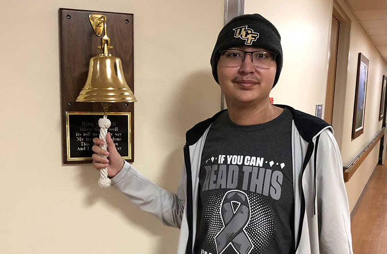 A young man in a black shirt and wool UCF cap rings a gold bell mounted on the wall.