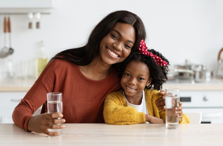 A young Black woman with her hair parted down the middle wearing a brown shirt is smiling as she sits at a table in her kitchen with a Black child wearing a white T-shirt, a mustard color sweater and a red bow with white polka dots in her hair who is also smiling. The background shows their kitchen blurred. They both have their hands wrapped around a glass of water sitting in front of them. 