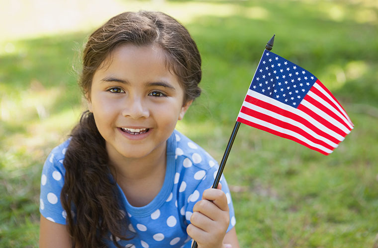 Little girl with brown hair and wearing blue shirt with white polka dots is holding a small American flag and smiling.