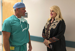 A man dressed in hospital scrubs and a head cover used in operating rooms smiles as he stands in a hospital corridor and talks with a woman with blond hair who may be a patient.