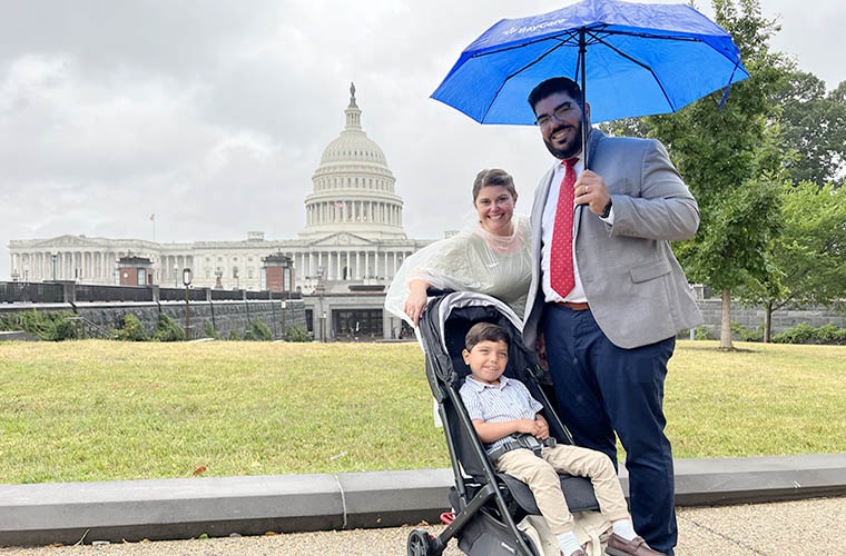 A man, woman and toddler who are a family are standing with a view of the U.S. Capitol building in the background. The toddler is a boy and is in a stroller. It is sprinkling and the mother is wearing a poncho and father is holding an umbrella. 