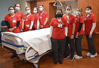 Nine students wearing face masks and red scrubs gather around a hospital bed.