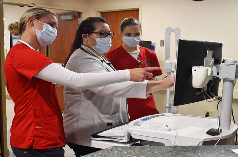 Students wearing face masks and red scrubs stand on either side of a woman wearing a white clinician's jacket as they discuss images they are viewing on a monitor. 