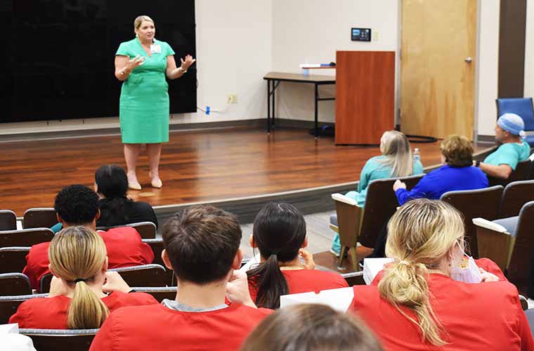 Woman wearing green dress addresses auditorium of filled with people wearing scrubs.