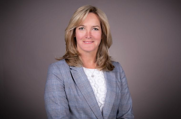 A headshot of Patricia Shucoski smiling wearing a gray windowpane-style suit jacket with a white lace top underneath that has a repeated circular design on it. She is wearing a pendant style necklace and has shoulder-length blonde hair with layers and parted on the left side.