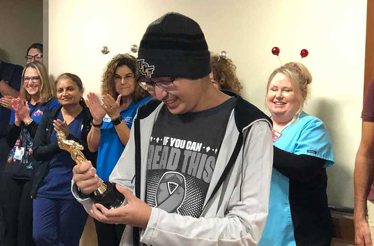 Aiya in a black shirt and wool UCF cap holds and looks smiling at a replica Oscar trophy.