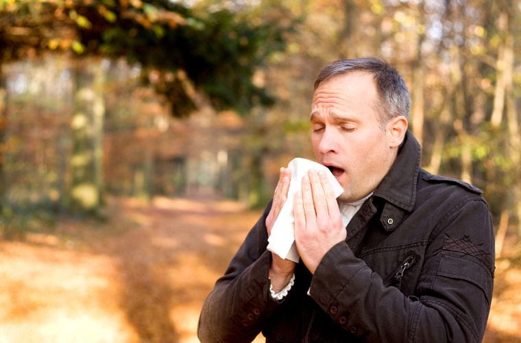 A middle-aged man with short salt and pepper hair and a receding hairline sneezes into a tissue standing in a nature scene. 