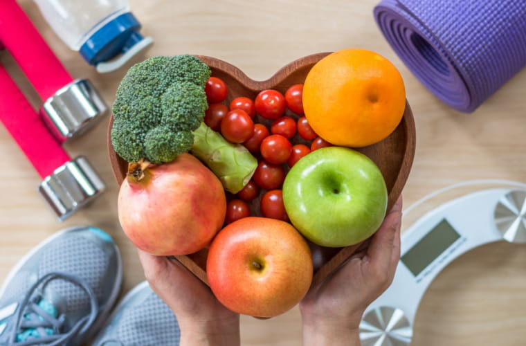 A person surrounded by workout gear holds a bowl of fresh fruit and vegetables.