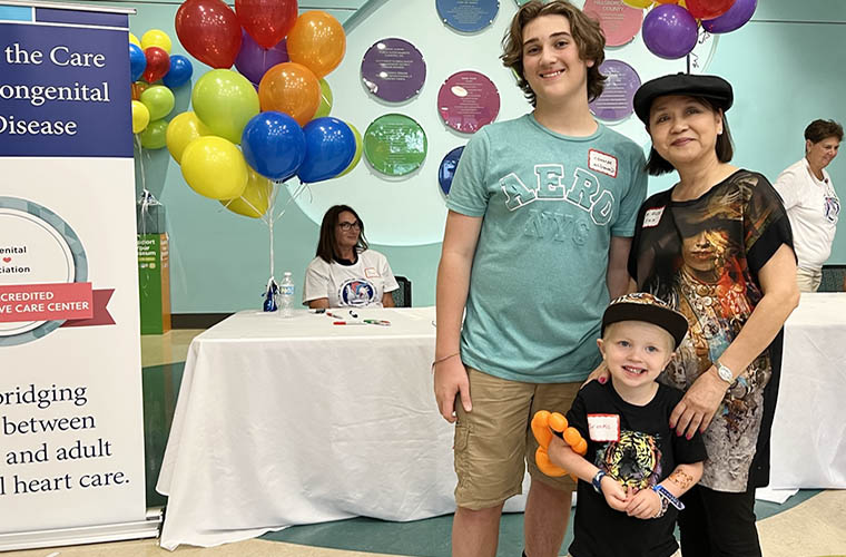 Teen and toddler boys smiling and standing next to one of their cardiologists, a woman. They are all having fun at a heart reunion event. 
