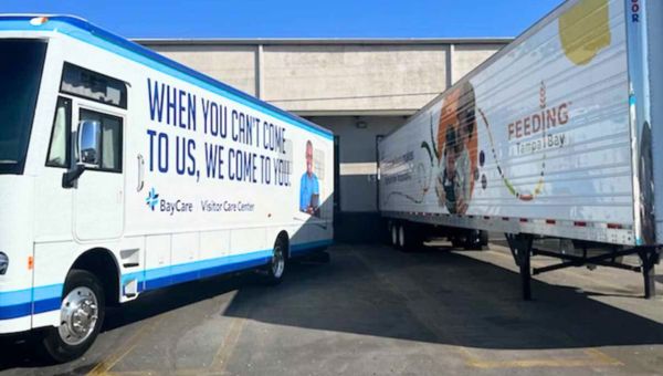 Two trailers, one representing BayCare and one representing Feeding Tampa Bay, are parked next to each other outside of a warehouse. The one on the left says, "When you can't come to us, we come to you," and the one on the right says, "Feeding Tampa Bay."