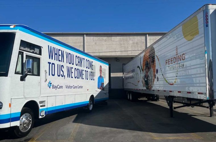 Two trailers, one representing BayCare and one representing Feeding Tampa Bay, are parked next to each other outside of a warehouse. The one on the left says, "When you can't come to us, we come to you," and the one on the right says, "Feeding Tampa Bay."