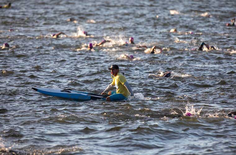 A man in a kayak sits in the water as swimmers glide past him in the water during a competition.