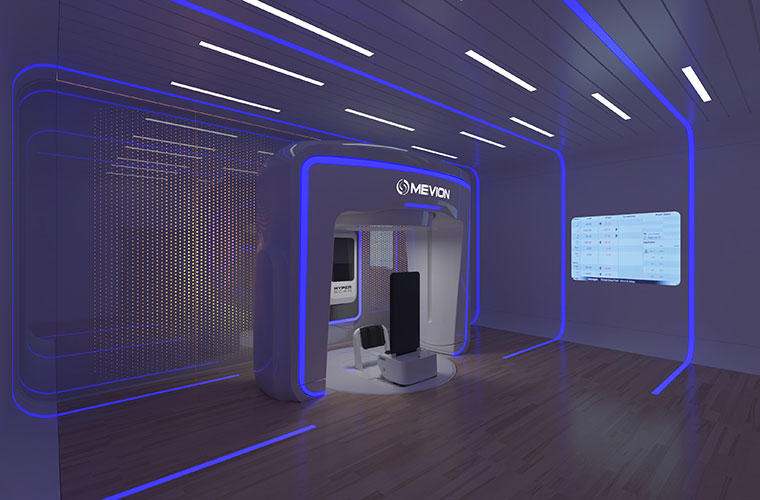The new proton therapy system in an open room. There is an upright area meant for patients to stand in while surrounded by the open large square. There are blue lights that accent the equipment.