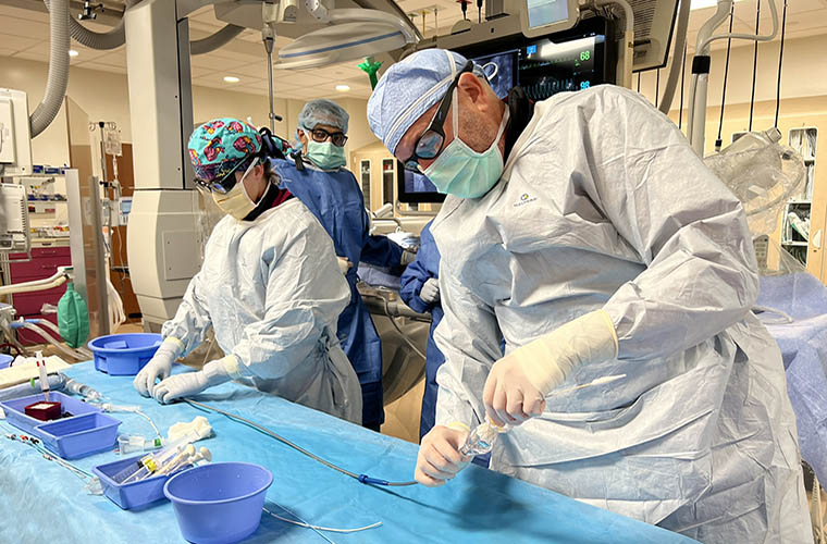 Three clinicians are in a hospital cardiac catherization laboratory and all are wearing surgical gowns, masks and caps. A woman is on the left and a male is on the right and they are handling medical equipment. The third person is in the background and watching what the other two are doing. 