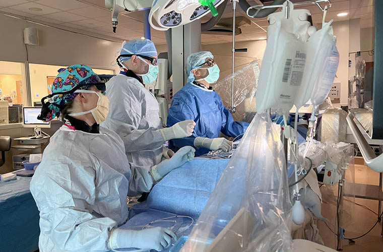 Three clinicians are in a hospital cardiac catherization laboratory and all are wearing surgical gowns, masks and caps. They are standing in front of a surgical table looking at the images on a large monitor.