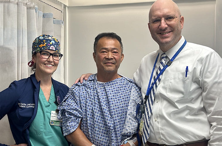 Three people smile at camera inside a hospital room. A woman is on the left and is wearing green scrubs and a surgical cap. A male hospital patient is in the middle and he is wearing a hospital gown. A male doctor is on the right and is wearing a white long-sleeve dress shirt.