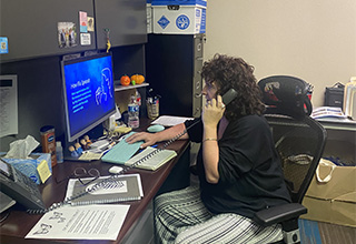 Woman with brown hair in a black top sitting in front of a computer screen at a desk talking on the phone.