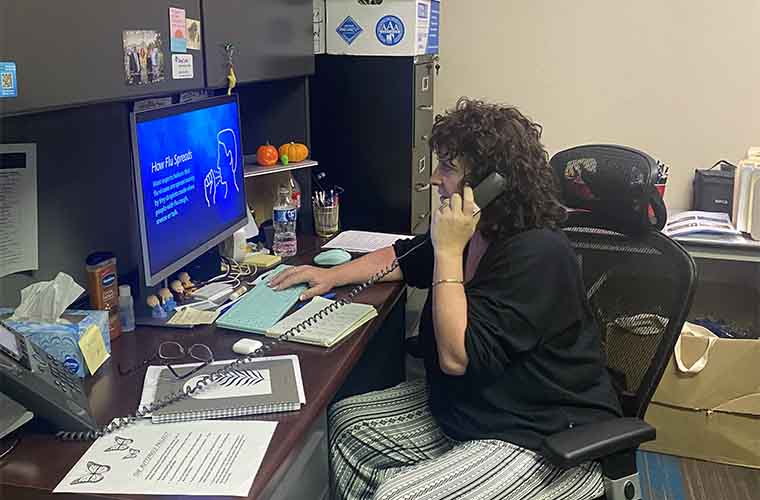 Woman with brown hair in a black top sitting in front of a computer screen at a desk talking on the phone.