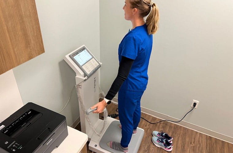 A woman dressed in hospital scrubs stands on a device that looks like a scale while holding controllers in her hands to demonstrate how the device will work.